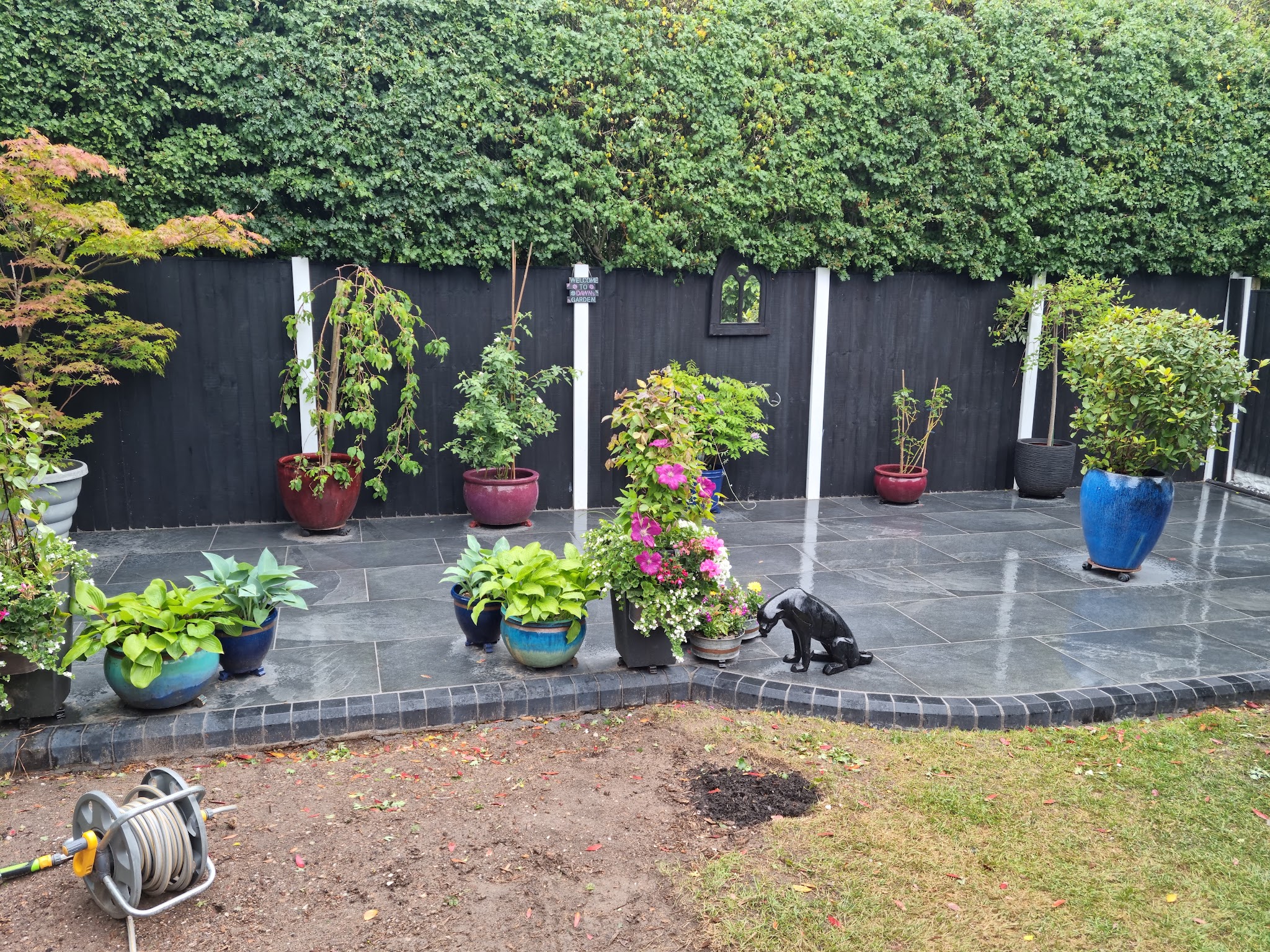 Dark porcelain patio with curved tiled edging, planted pots and a black fence behind